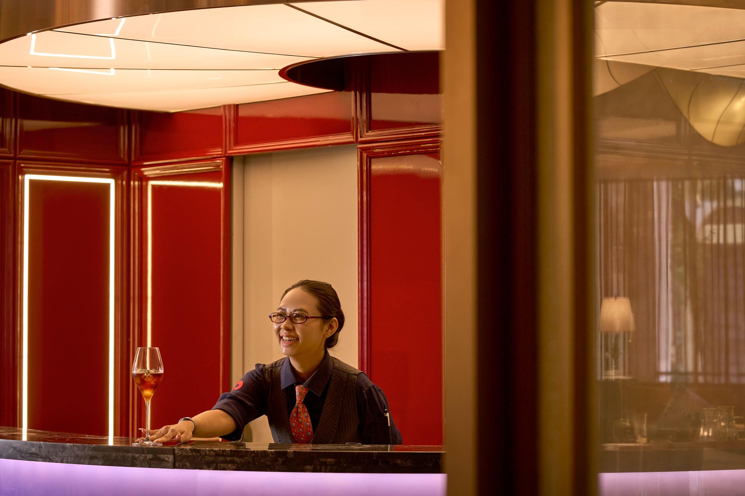 A barista smiles behind the curved bar, red lacquer panels and a curved ceiling light behind.