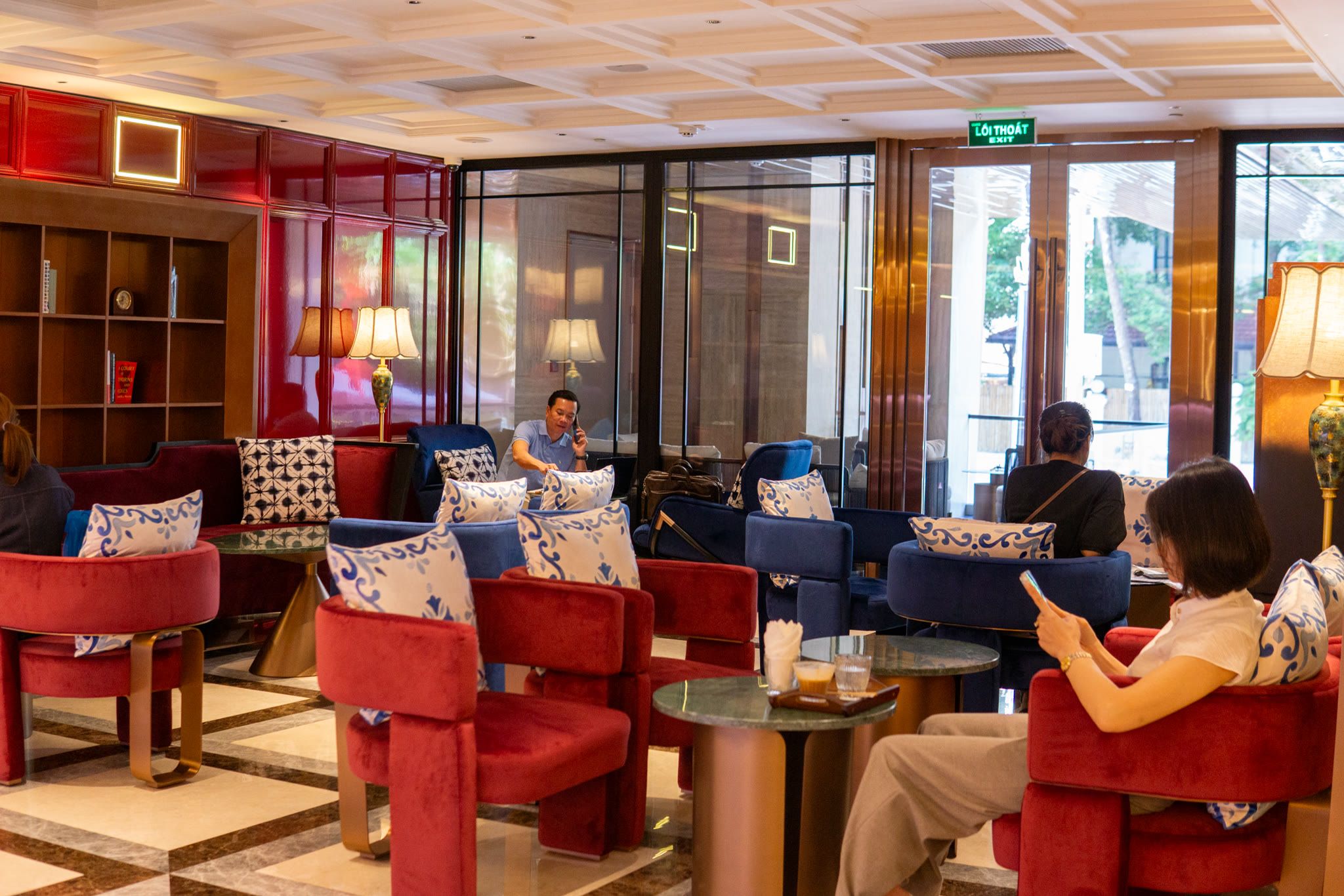 Guests at round stone tables between red velvet chairs, in daylight from the glass doors.