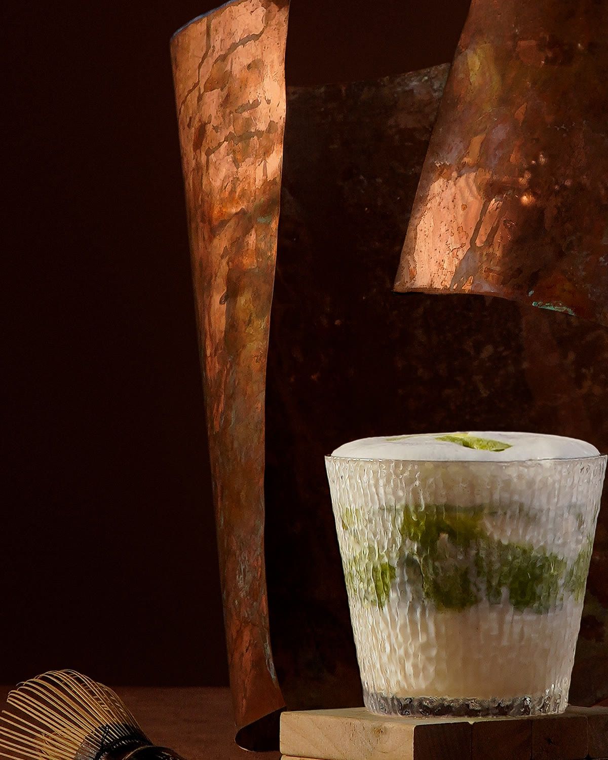 A glass of matcha latte topped with foam, a bamboo whisk beside it.