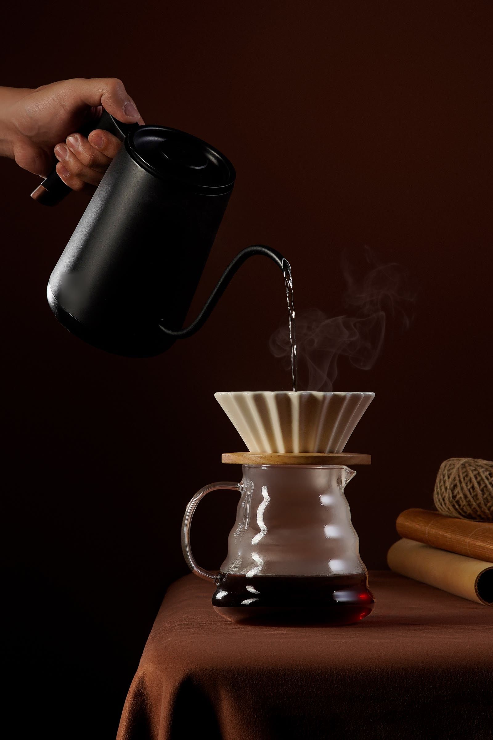 A hand pours water from a black gooseneck kettle into a ceramic dripper, steam rising in low, warm light.